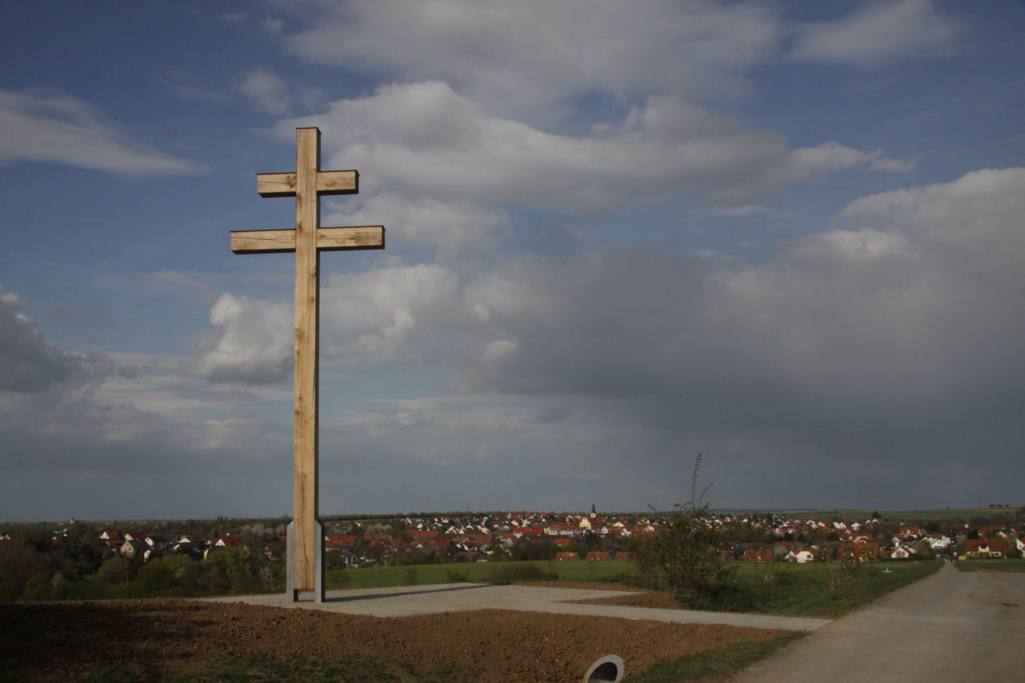 Blick über Kürnach  Ein Platz zum Verweilen, Ausruhen und Genießen Kürnach (Mad)  Im Herbst letzten Jahres hat die Ehrenbürgerin der Gemeinde Kürnach Christine Demel gemeinsam mit etlichen Vereinen, den Feldgeschworenen, Einzelpersonen und ihrer Familie ein Patriarchen Kreuz am Haflersweg anlässlich ihres 80. Geburtstages gestiftet und errichten lassen. Auf einer Anhöhe ragt es nun über Kürnach. Nun hat der Gemeinderat beschlossen, dort einen Platz zum Verweilen, Ausruhen und auch Genießen zu schaffen. Die Firma Ullrichbau gestaltet im Auftrag der Gemeinde eine kleine Pflasterfläche, auf der ein Tisch und zwei Bänke Platz finden werden, die von Schülern der Bauinnung Würzburg unter Leitung von Benedikt Scheller hergestellt worden sind. „Hier können Wanderer und Radler Brotzeit machen, über Kürnach blicken, bei guter Sicht gar bis in die Rhön, sich ausruhen oder einfach ein bisschen zum Nachdenken kommen“ freut sich Bürgermeister Thomas Eberth. „Heutzutage sind Plätze der Entspannung wichtiger denn je“ ist Eberth sicher, während sich Christine Demel besonders darüber freut, dass das Kreuz von den Einwohnern Kürnachs derart positiv angenommen wird. Ein Schild wird das Patriarchen Kreuz näher erläutern. Der obere, kürzere Querbalken symbolisiert die Inschrift INRI auf dem Kreuz Jesu Christi, dem lateinischen Kreuz. Derartige Kreuze wurden im 9. Jh. von den griechischen Heiligen und Gelehrten Kyrill und Method nach Ungarn gebracht. Bald standen sie überall in Europa als Schutzschilde gegen Unwetter, Blitz, Krankheiten und kriegerische Ereignisse an Ortsverbindungsstraßen um die Siedlungen und bis heute auf Kirchengebäuden. Der Name Patriarchenkreuz erinnert an die Patriarchen von Jerusalem, denen ein derartiges Kreuz vorausgetragen wurde. Kyrill und Method wurden durch Papst Johannes Paul II. im Jahr 1980 Schutzpatrone Europas.  Bild: Bürgermeister Thomas Eberth (rechts) erläutert zusammen mit Ehrenbürgerin Demel den Bauarbeitern Polier Hartwig Müller (links) Manfred Haupt (2.v.l.) und Baggerfahrer Fred Ehrenberg die Gestaltung des Platzes.  